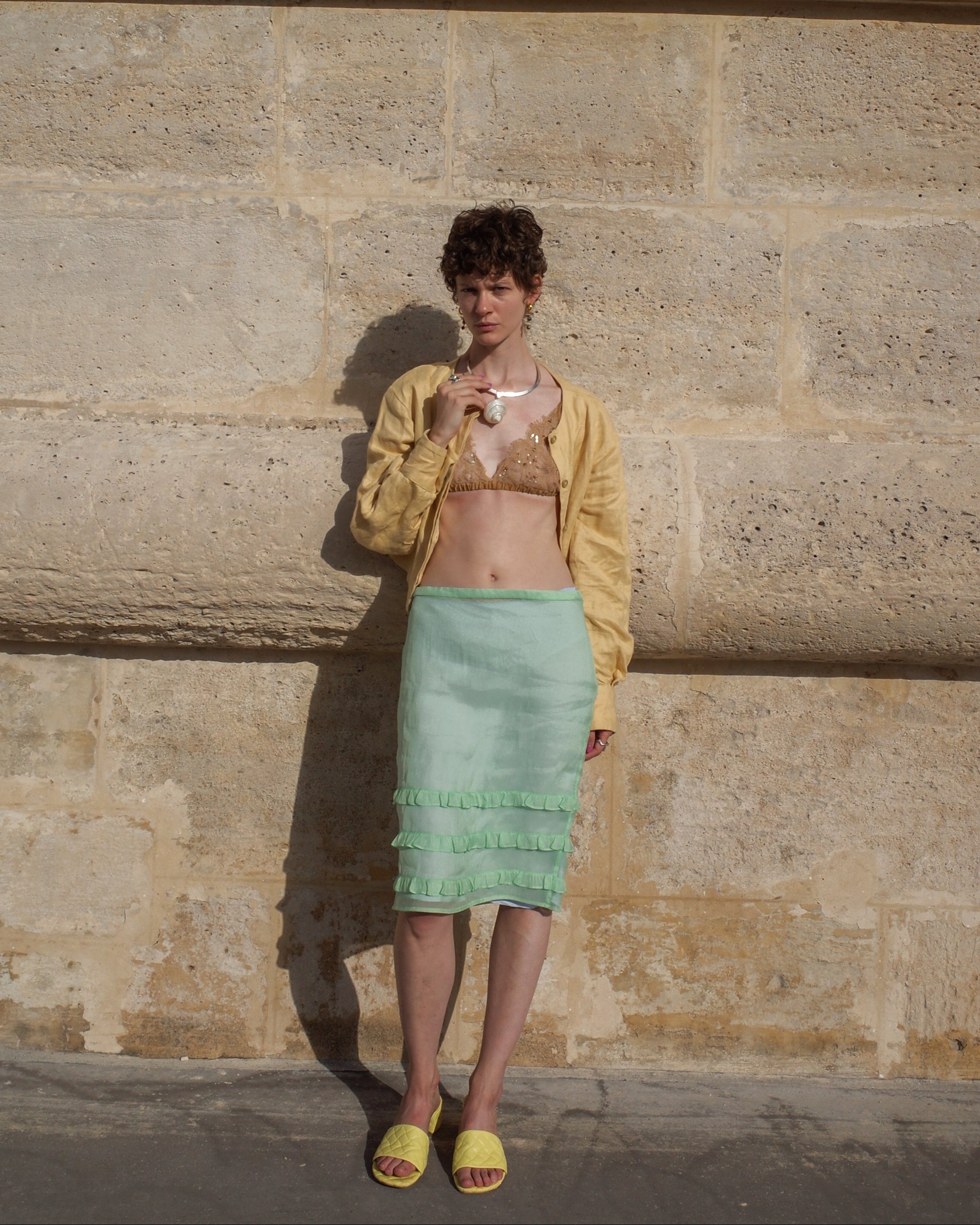 Woman in yellow jacket and green skirt standing against a stone wall.