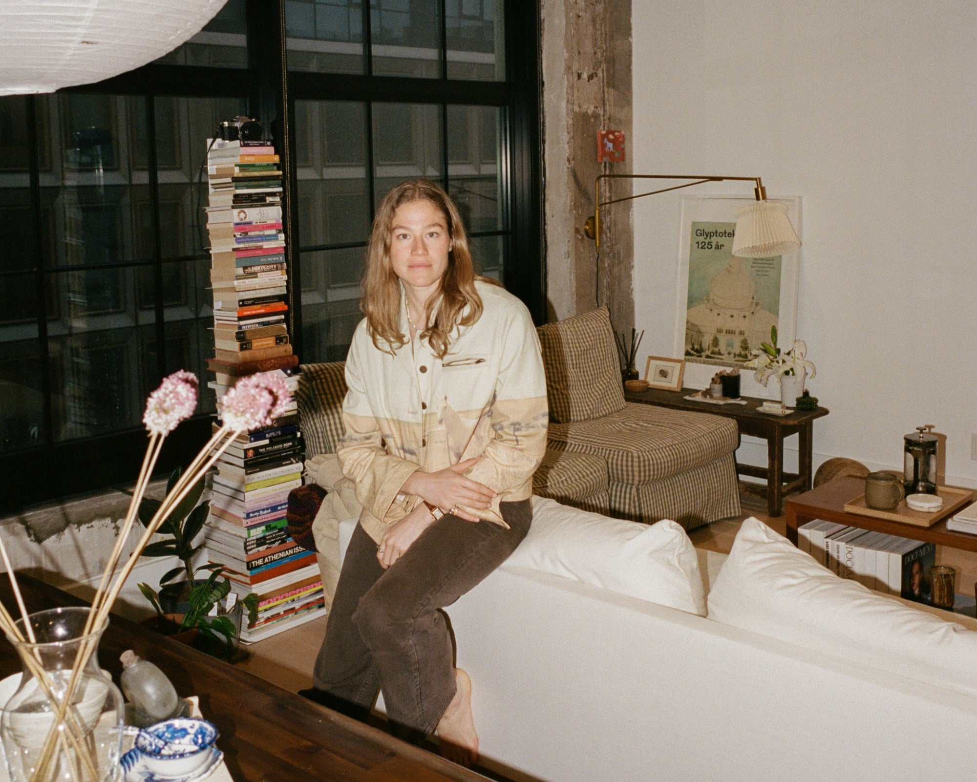 Woman sitting on a couch in a modern living room with large windows and decorative elements.